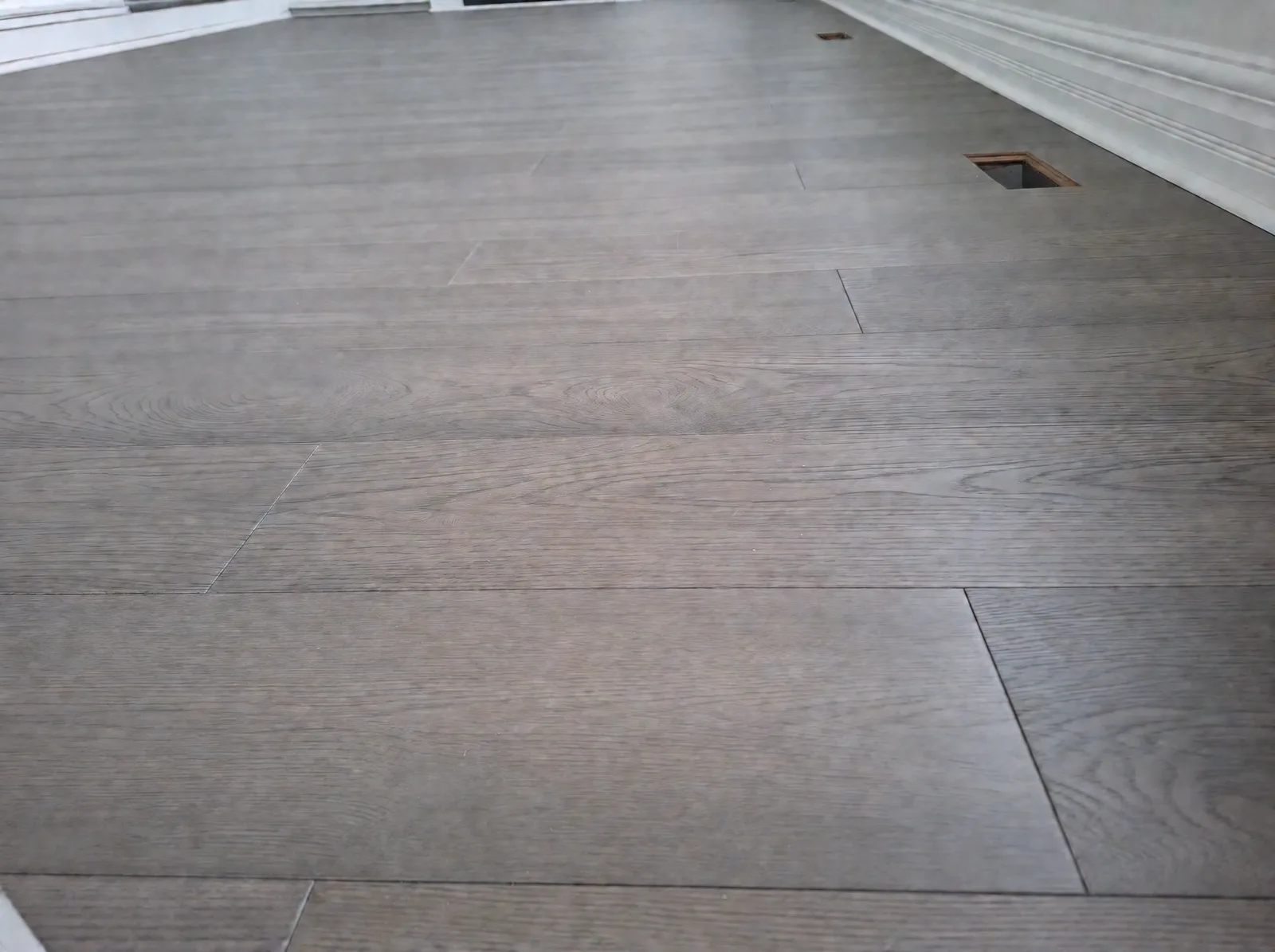 Close-up of wire-brushed grey oak planks with a floor register cut-out and white baseboard