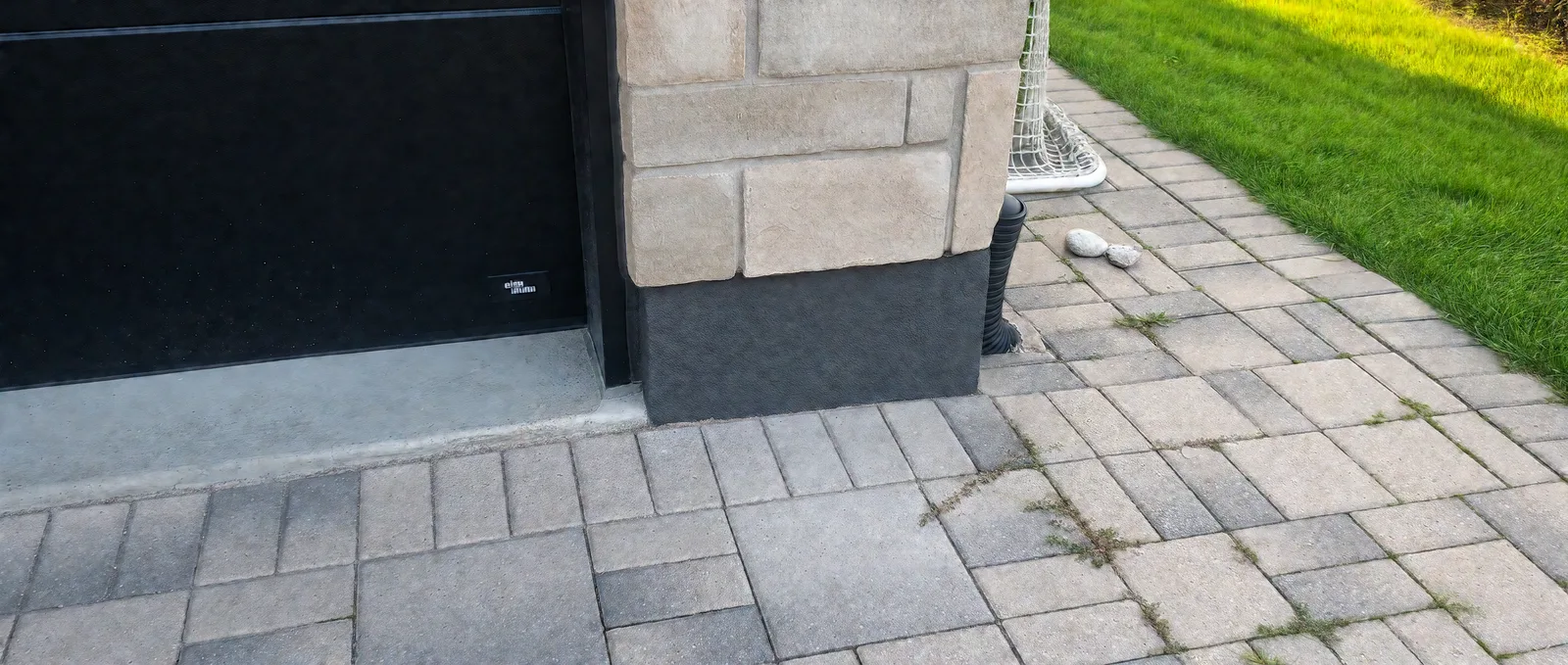 Finished charcoal parging beneath stone veneer at a garage corner, beside the black garage door and paver driveway